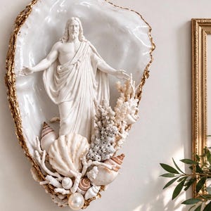 May include: A wall-mounted decorative piece featuring a white resin figure of Jesus with arms outstretched, set inside a large, iridescent oyster shell with gold-painted edges. The base is adorned with various white seashells, coral fragments, and a single pearl.