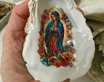 Puede incluir: Una concha de ostra blanca con una imagen religiosa de una mujer con una túnica azul, rodeada de flores. La concha se sostiene en una mano, con otras conchas en el fondo.