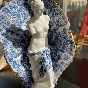 Pu&ograve; includere: Una statua in gesso bianco di una donna in piedi in un guscio di porcellana blu e bianco. Il guscio ha un bordo dorato.