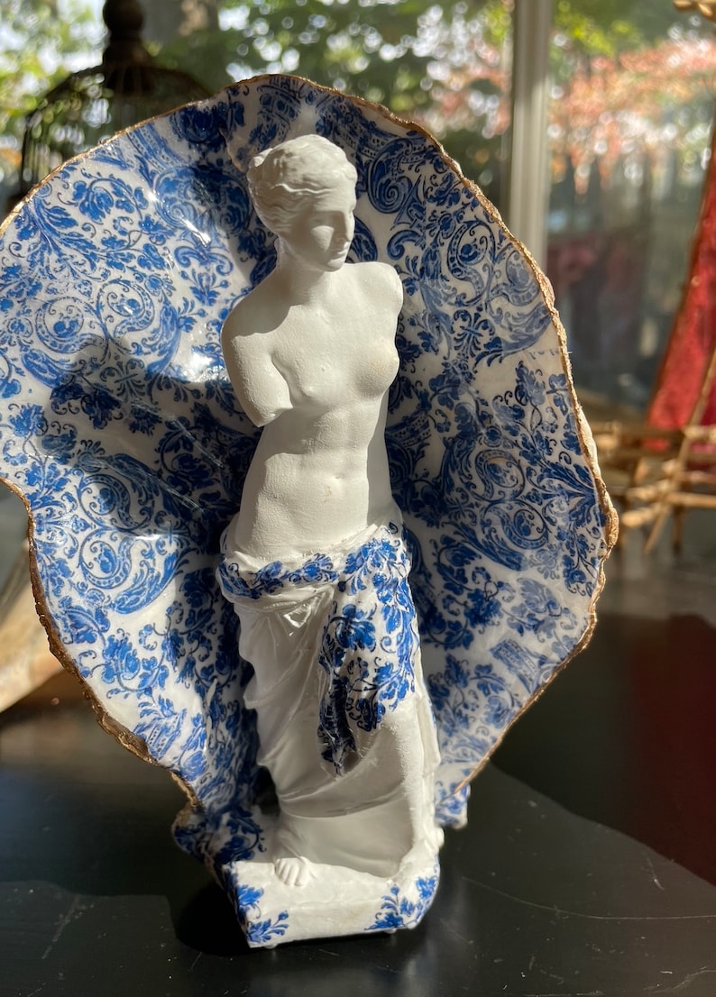Pu&ograve; includere: Una statua in gesso bianco di una donna in piedi in un guscio di porcellana blu e bianco. Il guscio ha un bordo dorato.