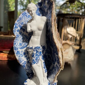 Pu&ograve; includere: Una statua in porcellana bianca di una donna in piedi in un guscio di porcellana blu e bianco. Il guscio ha un bordo dorato.
