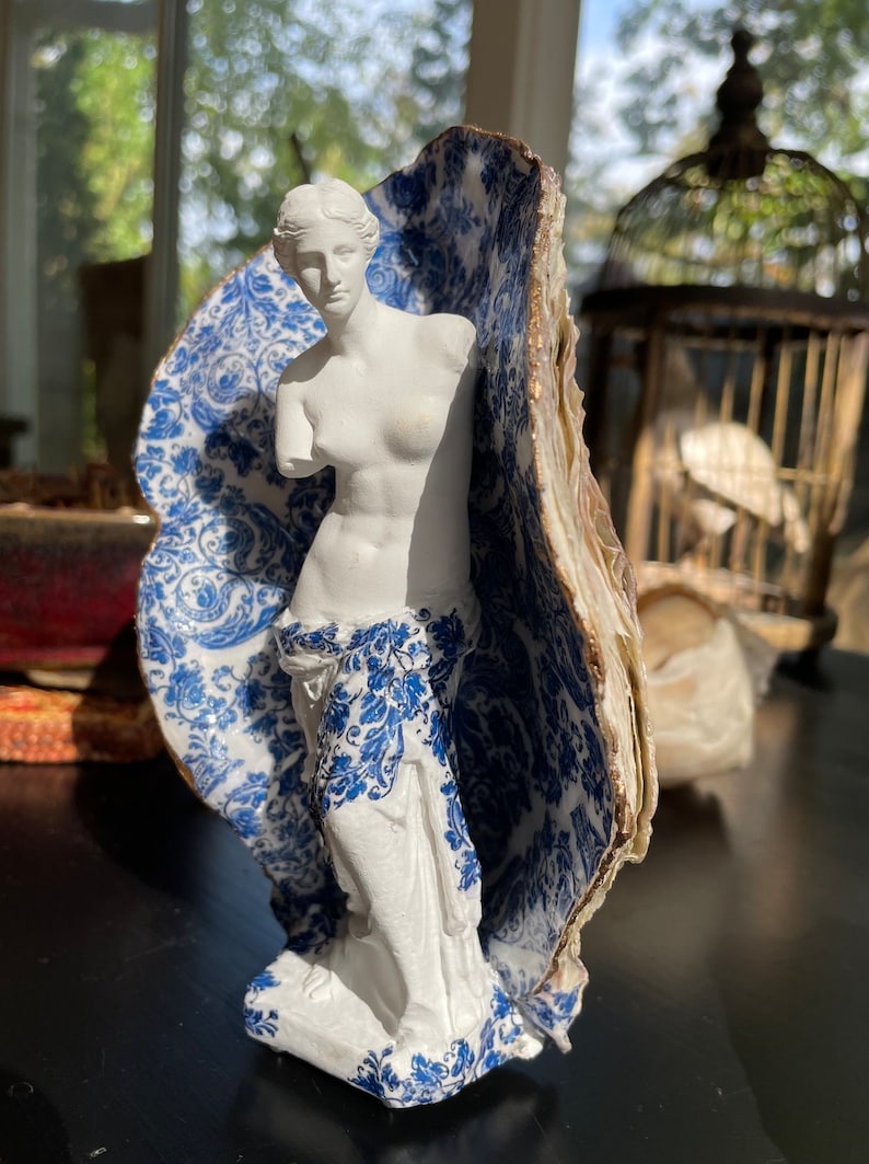 Pu&ograve; includere: Una statua in porcellana bianca di una donna in piedi in un guscio di porcellana blu e bianco. Il guscio ha un bordo dorato.