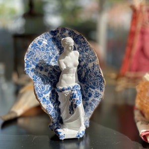Pu&ograve; includere: Una statua in porcellana bianca di una donna in piedi con un tessuto drappeggiato su di lei con un motivo blu e bianco. Il tessuto ha un motivo floreale.