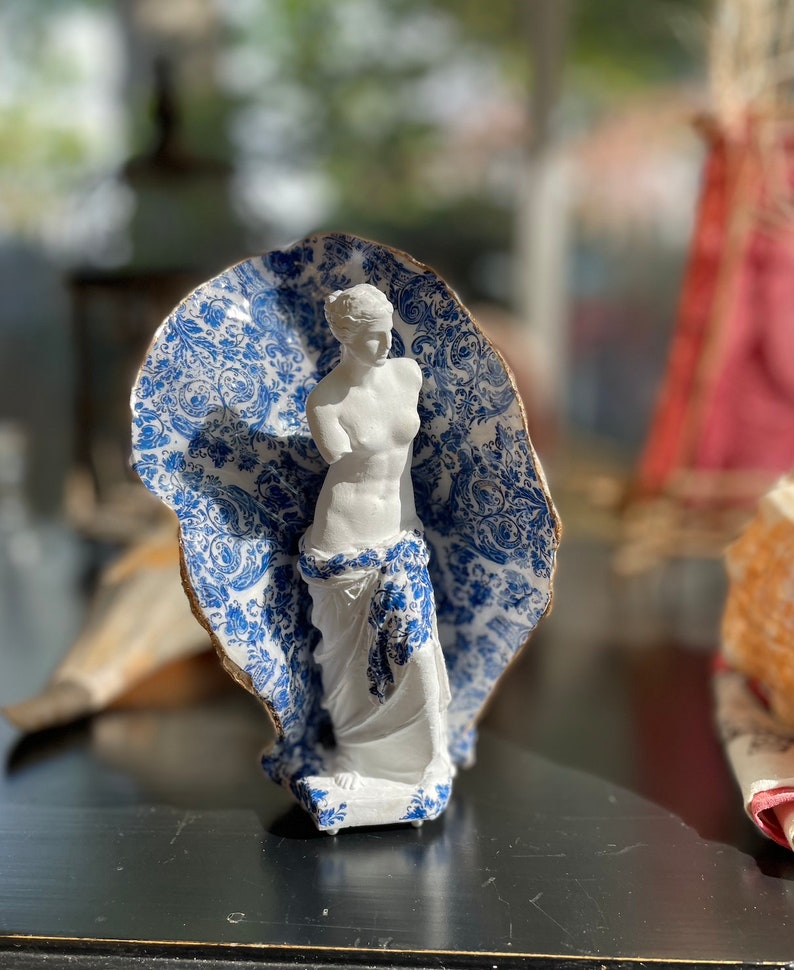 Pu&ograve; includere: Una statua in porcellana bianca di una donna in piedi con un tessuto drappeggiato su di lei con un motivo blu e bianco. Il tessuto ha un motivo floreale.