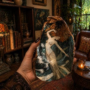 Puede incluir: Una pieza de arte en concha pintada a mano con una sirena. El interior de la concha muestra una sirena con cabello suelto y una cola brillante, sobre un fondo de olas oceánicas. Los bordes de la concha están adornados con oro.