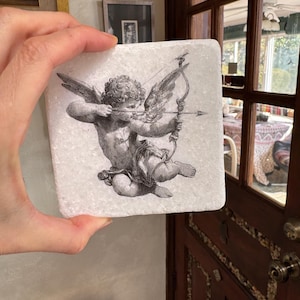 May include: Square white marble coaster featuring a detailed black and white illustration of Cupid, the Roman god of love, aiming a bow and arrow. The artwork has a vintage, classical aesthetic. The coaster is held up by a hand.