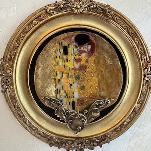 Könnte beinhalten: Ein rundes, goldfarben gerahmtes Kunstwerk mit Gustav Klimts "Der Kuss". Das Kunstwerk zeigt ein sich umarmendes Paar mit aufwendigen Gold- und Gelbdetails. Der Rahmen hat eine verzierte Gestaltung und einen dunklen Innenrand.
