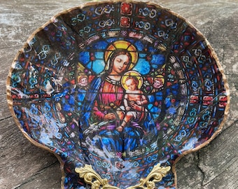 Puede incluir: Un plato decorativo en forma de concha de vieira con una imagen de vitral de la Virgen María y el niño Jesús. La concha está pintada con un borde dorado y tiene un asa de metal dorado.
