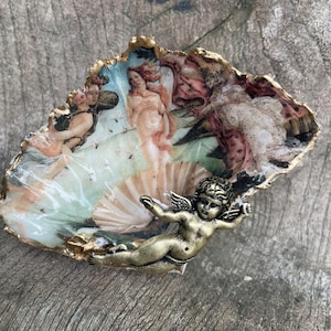 K&ouml;nnte beinhalten: Dekorative Austernschale mit einem Druck von Botticellis "Die Geburt der Venus". Die Schale hat einen goldfarbenen Rand und eine kleine Bronze-Cherubfigur. Das Kunstwerk zeigt klassische Figuren in sanften Farben.