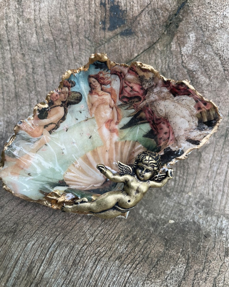 K&ouml;nnte beinhalten: Dekorative Austernschale mit einem Druck von Botticellis "Die Geburt der Venus". Die Schale hat einen goldfarbenen Rand und eine kleine Bronze-Cherubfigur. Das Kunstwerk zeigt klassische Figuren in sanften Farben.