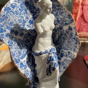 Pu&ograve; includere: Una statua di porcellana bianca di una donna, in piedi su una base con un motivo blu e bianco. La statua &egrave; parzialmente coperta da un guscio con un motivo blu e bianco.