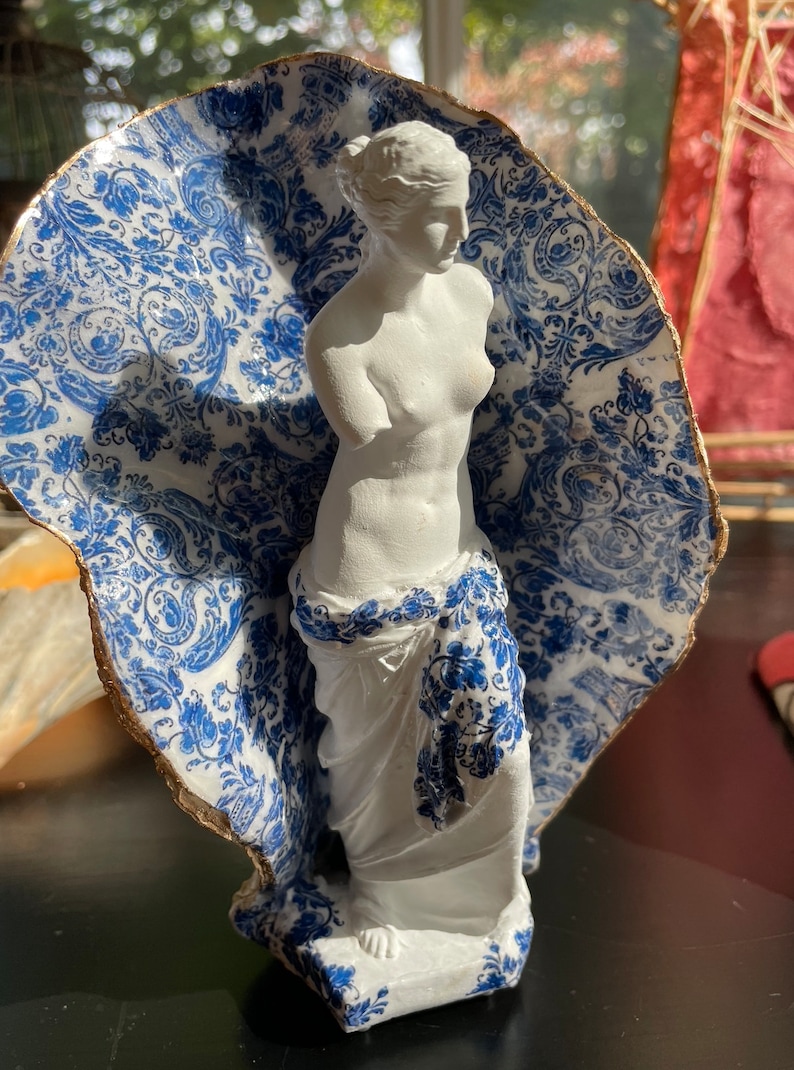 Pu&ograve; includere: Una statua di porcellana bianca di una donna, in piedi su una base con un motivo blu e bianco. La statua &egrave; parzialmente coperta da un guscio con un motivo blu e bianco.