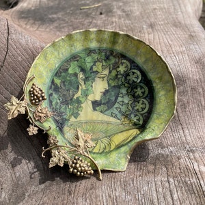 May include: A decorative, shell-shaped dish in green and gold. The dish features a central image of a woman with ivy in her hair. A gold-coloured grape vine and grapes adorn the side. The dish is placed on a wooden surface.