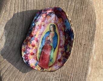 Puede incluir: Un plato redondo decorativo de colores con una imagen de la Virgen María rodeada de flores. El plato está hecho de un material natural, posiblemente madera o concha.