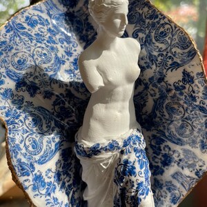 Pu&ograve; includere: Una statua in gesso bianco della Venere di Milo, in piedi su una base in ceramica con motivo blu e bianco. La base ha la forma di una conchiglia e un bordo dorato.
