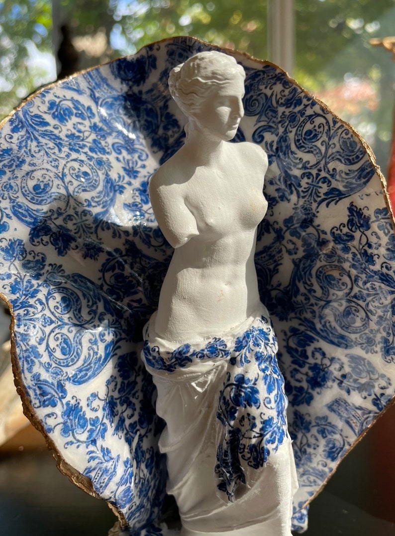 Pu&ograve; includere: Una statua in gesso bianco della Venere di Milo, in piedi su una base in ceramica con motivo blu e bianco. La base ha la forma di una conchiglia e un bordo dorato.