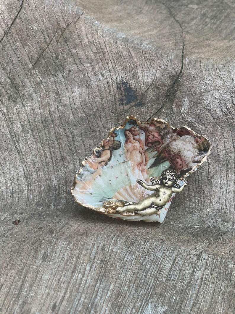 K&ouml;nnte beinhalten: Eine dekorative, muschelf&ouml;rmige Schale mit einer bemalten Szene klassischer Figuren und einem goldfarbenen Rand. Eine kleine, goldfarbene Cherubfigur ist an der Schale befestigt. Die Schale liegt auf einer verwitterten, grauen Holzoberfl&auml;che.