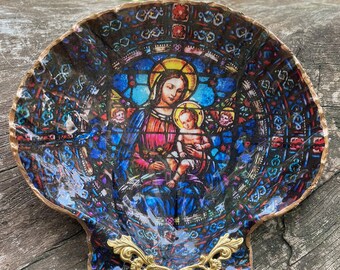 Puede incluir: Un plato decorativo en forma de concha de mar con una imagen de vitral de la Virgen María y el niño Jesús. La concha está pintada con un borde dorado y tiene un asa de metal dorado.