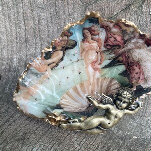 K&ouml;nnte beinhalten: Dekorative Austernschale mit einem Renaissance-Gem&auml;lde der Venus. Die Schale hat einen goldfarbenen Rand und einen kleinen, bronzefarbenen Putto. Das Kunstwerk zeigt Figuren in Rosa-, Pfirsich- und Blaut&ouml;nen.