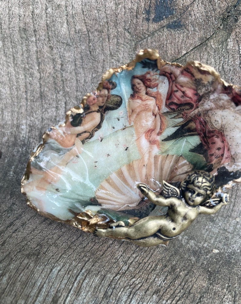 K&ouml;nnte beinhalten: Dekorative Austernschale mit einem Renaissance-Gem&auml;lde der Venus. Die Schale hat einen goldfarbenen Rand und einen kleinen, bronzefarbenen Putto. Das Kunstwerk zeigt Figuren in Rosa-, Pfirsich- und Blaut&ouml;nen.