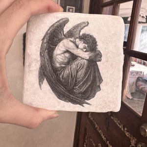 以下が含まれることがあります： オフホワイトの正方形の石コースター。黒と白で描かれた翼のある人物のイラストが特徴です。その人物はひざの上に頭を乗せて座っており、暗い布に覆われ、翼を広げています。