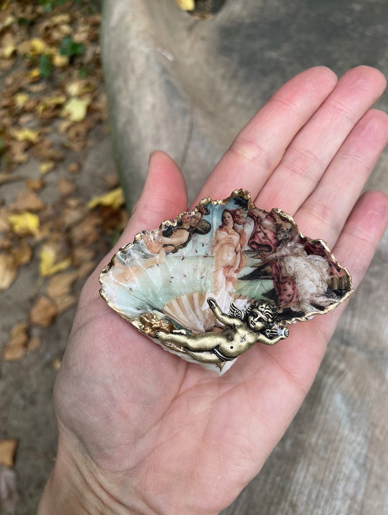 K&ouml;nnte beinhalten: Eine dekorative, muschelf&ouml;rmige Schmuckschale mit einem Druck von Botticellis "Die Geburt der Venus". Die Schale hat einen goldfarbenen Rand und eine kleine, goldfarbene Cherubfigur. Die Muschel wird in einer Hand gehalten.