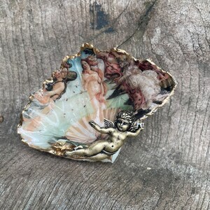 K&ouml;nnte beinhalten: Eine muschelf&ouml;rmige dekorative Schale mit goldfarbenem Rand und einem gemalten Bild eines klassischen Kunstwerks. Eine kleine, bronzefarbene Cherubfigur ist an der Schale befestigt. Die Schale liegt auf einer verwitterten Holzoberfl&auml;che.