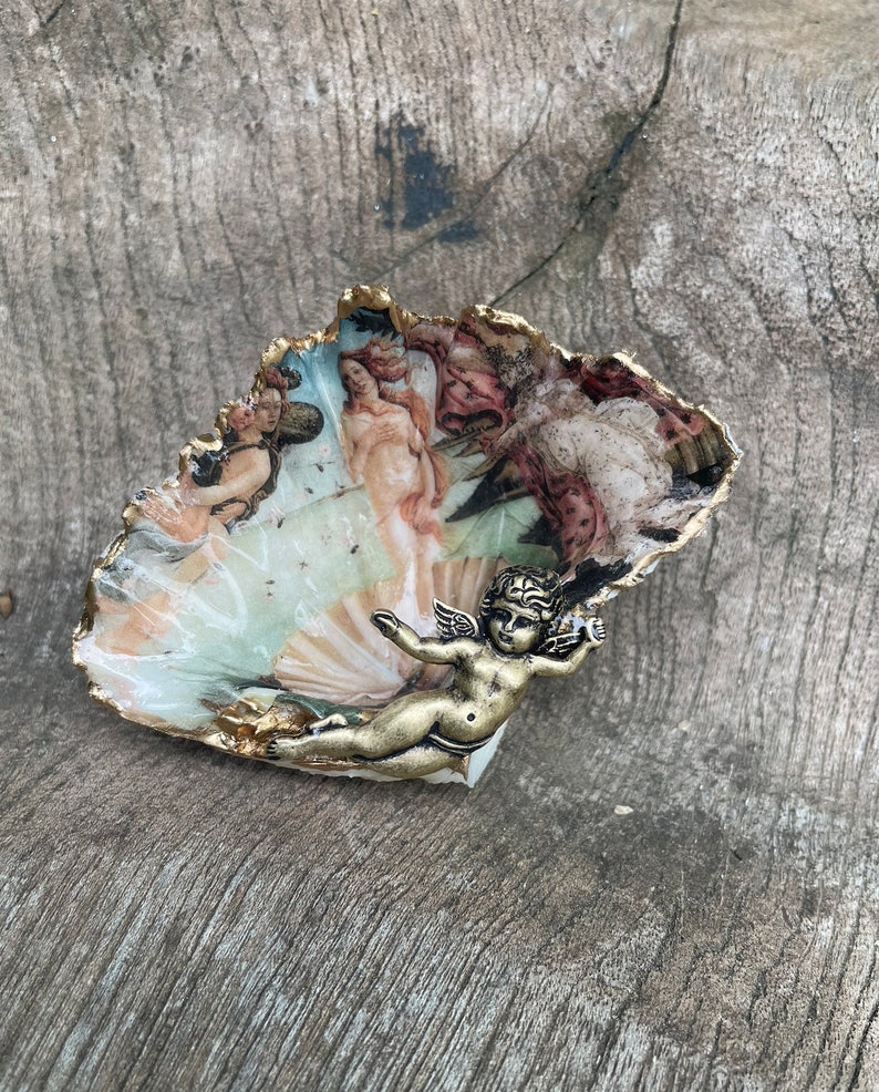 K&ouml;nnte beinhalten: Eine muschelf&ouml;rmige dekorative Schale mit goldfarbenem Rand und einem gemalten Bild eines klassischen Kunstwerks. Eine kleine, bronzefarbene Cherubfigur ist an der Schale befestigt. Die Schale liegt auf einer verwitterten Holzoberfl&auml;che.