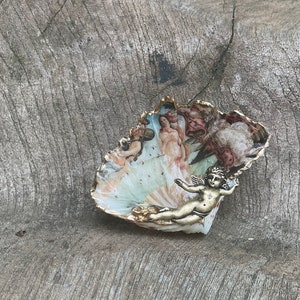 K&ouml;nnte beinhalten: Eine dekorative, muschelf&ouml;rmige Schale mit einer bemalten Szene klassischer Figuren und einem goldfarbenen Rand. Eine kleine, goldfarbene Cherubfigur ist an der Schale befestigt. Die Schale liegt auf einer verwitterten, grauen Holzoberfl&auml;che.