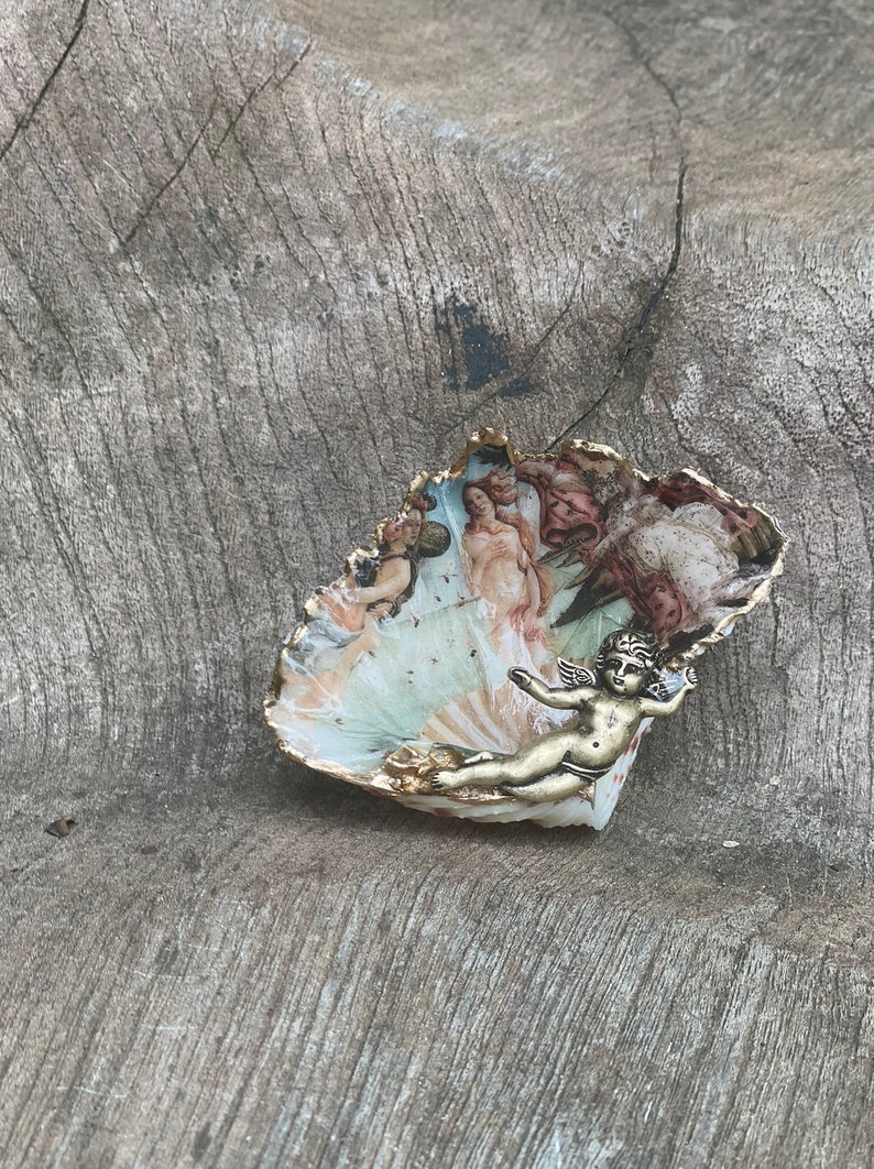 K&ouml;nnte beinhalten: Eine dekorative, muschelf&ouml;rmige Schale mit einer bemalten Szene klassischer Figuren und einem goldfarbenen Rand. Eine kleine, goldfarbene Cherubfigur ist an der Schale befestigt. Die Schale liegt auf einer verwitterten, grauen Holzoberfl&auml;che.