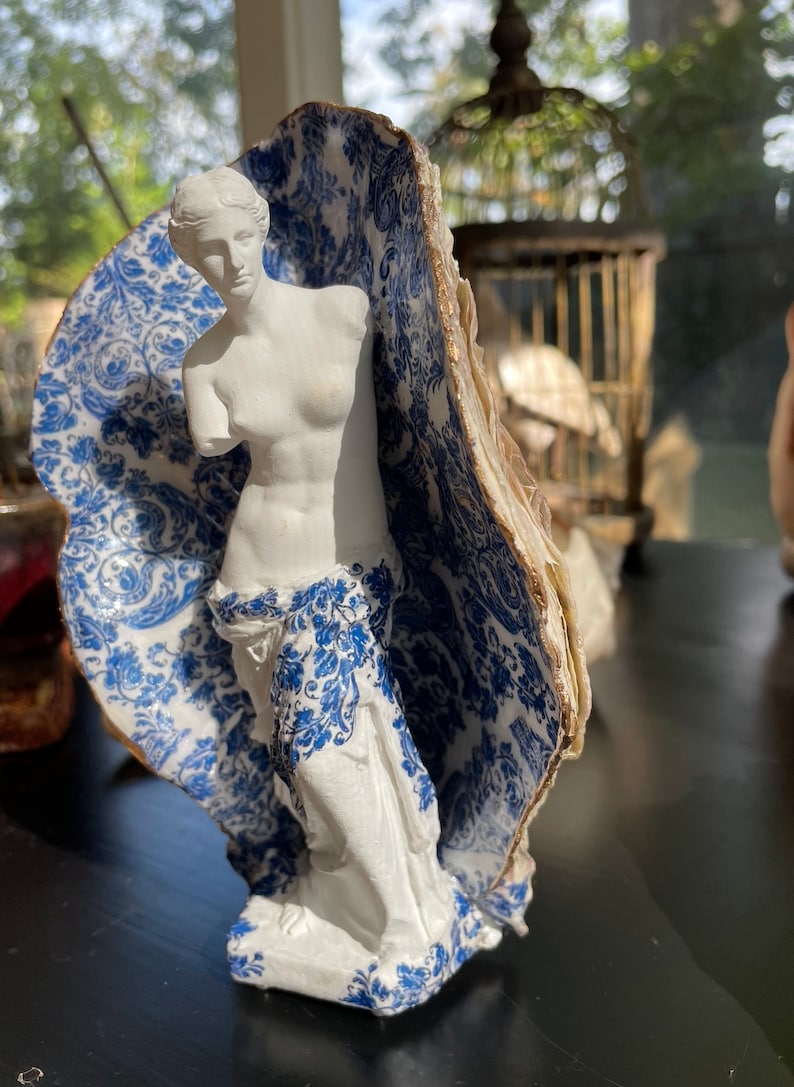 Pu&ograve; includere: Una statua in gesso bianco di una donna, in piedi in un guscio di ostrica con motivo blu e bianco. La statua &egrave; una replica della Venere di Milo.