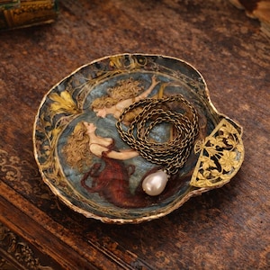 Puede incluir: Un plato decorativo en forma de concha con una escena de sirenas pintadas y detalles dorados. Un collar de cadena dorada con un colgante de perla descansa dentro. El plato está sobre una superficie de madera oscura.