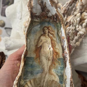 May include: A decorative oyster shell featuring a painted image of Botticelli's "The Birth of Venus." The shell is trimmed with gold paint and adorned with floral and seashell accents. The artwork depicts Venus standing in a shell, with long flowing red hair.