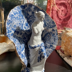 Pu&ograve; includere: Una statua di porcellana bianca di una donna, parzialmente coperta da un guscio di ceramica con motivi blu e bianchi. Il guscio ha un bordo dorato.