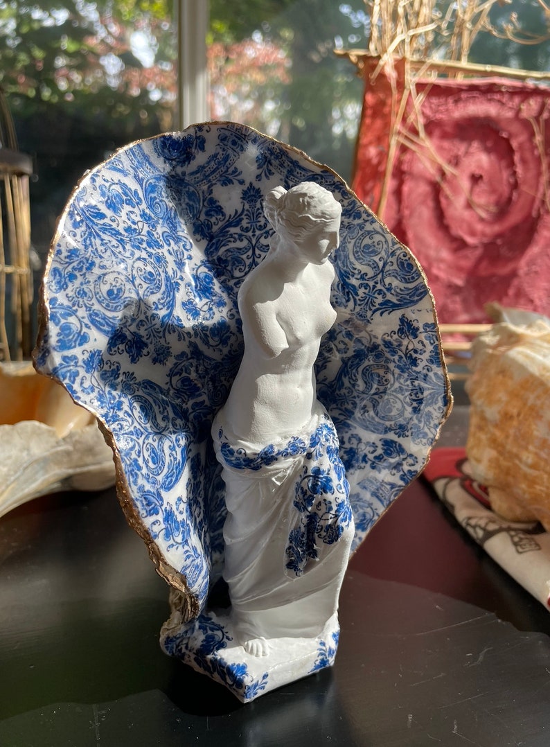 Pu&ograve; includere: Una statua di porcellana bianca di una donna, parzialmente coperta da un guscio di ceramica con motivi blu e bianchi. Il guscio ha un bordo dorato.