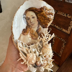 Può includere: Una conchiglia bianca con un dipinto di un viso di donna. La conchiglia è decorata con piccole conchiglie, coralli e perle.