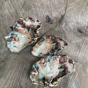 K&ouml;nnte beinhalten: Drei dekorative, muschelf&ouml;rmige Schalen mit einem Renaissance-Gem&auml;ldedesign. Die R&auml;nder sind goldfarben verziert, und jede Schale hat eine kleine Bronzefigur eines Engels. Die Schalen stehen auf einer verwitterten Holzoberfl&auml;che.
