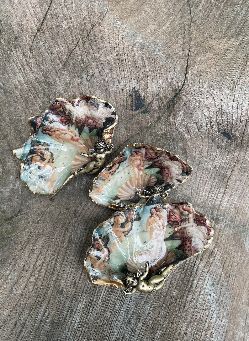 K&ouml;nnte beinhalten: Drei dekorative, muschelf&ouml;rmige Schalen mit einem Renaissance-Gem&auml;ldedesign. Die R&auml;nder sind goldfarben verziert, und jede Schale hat eine kleine Bronzefigur eines Engels. Die Schalen stehen auf einer verwitterten Holzoberfl&auml;che.
