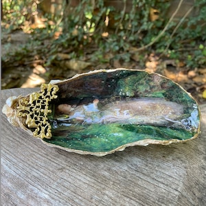 May include: A decorative oyster shell with a gold filigree accent. Inside the shell is a painting of a woman lying in a green meadow.