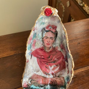 Puede incluir: Una concha de ostra pintada con un retrato de Frida Kahlo en su interior. La concha está decorada con flores y una rosa roja en la parte superior.