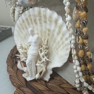 May include: A white sculpture of Venus stands before a large, cream-colored seashell. The sculpture is surrounded by white coral and seashells. Strands of seashells and shell beads hang nearby, with a woven, brown base.