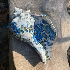 May include: A white seashell with a blue and white floral pattern. The shell is decorated with gold trim and has a scalloped edge.
