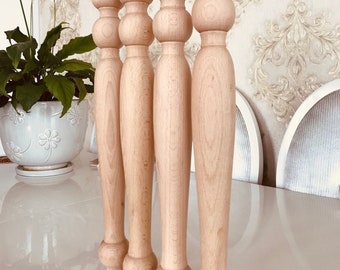 May include: Four unfinished wooden table legs with a turned design. The legs are arranged in a row, with the top of each leg facing the viewer. The legs are made of a light-colored wood and have a smooth finish.