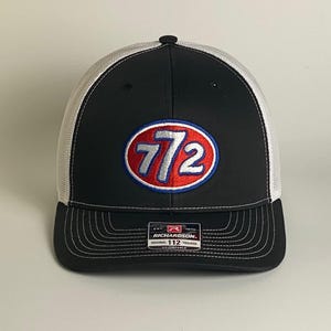 May include: Black and white trucker hat with a curved brim. The front panel features an embroidered oval logo with the number "772" in red and silver. The hat has a white mesh back and a black adjustable strap.