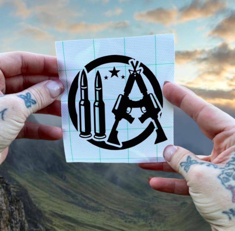 Puede incluir: Pegatina en blanco y negro con un dise&ntilde;o circular que presenta dos rifles cruzados en forma de "A", dos balas y una estrella. La pegatina est&aacute; sobre un fondo blanco.