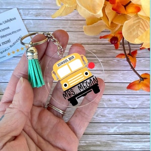 May include: A round, clear acrylic keychain with a yellow school bus illustration and the text "SCHOOL BUS" and "MRS. MEGAN". The keychain has a green tassel attached to a silver key ring.