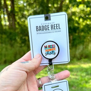 Puede incluir: Un carrete de identificación negro con una insignia redonda de colores que dice "Nurses Call the Shots". El carrete de identificación está unido a un porta-insignias de plástico transparente. El carrete de identificación está hecho a mano y es un porta-insignias intercambiable.