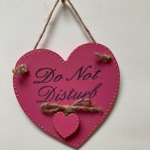 Könnte beinhalten: Ein herzförmiges Holzschild in Rosa mit dem Schriftzug "Do Not Disturb" in schwarzer Schrift. Es hat ein kleines Herz, goldene Punkte und eine Juteschnur zum Aufhängen. Ein dekorativer Artikel für eine Tür oder Wand.
