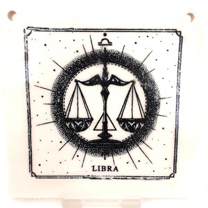 May include: Square, white tile with a black Libra zodiac symbol. The design features scales, a dotted circle, and radiating lines. The word "LIBRA" is at the bottom. The tile has two small holes at the top for hanging.