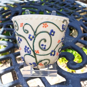 May include: A cream-coloured ceramic planter, decorated with blue and orange floral patterns and green vines. The rim has small, raised white dots. The planter is displayed on a clear acrylic stand. The planter is suitable for small plants.